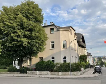 Ansicht eines Mehrfamilienhauses an einer Straßenecke mit altem Baumbestand und gepflegter Hecke.