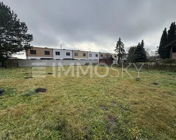 Leeres Grundstück mit Grasfläche und Bäumen, umgeben von einer Betonmauer und Wohnhäusern im Hintergrund.