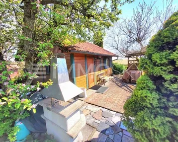 Gartenbereich mit Grillplatz und kleiner Holzhütte, umgeben von blühenden Bäumen und einer gepflasterten Terrasse.