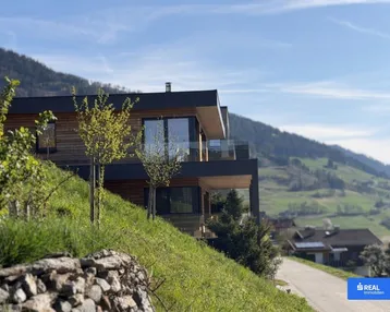 Modernes Haus in Hanglage mit Holzfassade, großen Fenstern und Panoramablick auf die Berge.