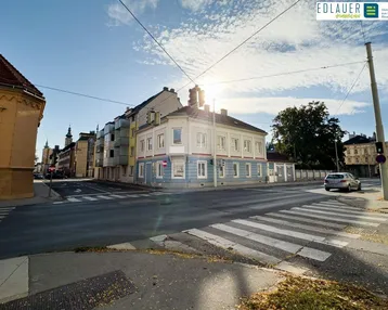 Das Eckgebäude präsentiert sich an einer belebten Kreuzung mit blauer und beiger Fassade unter strahlendem Himmel.