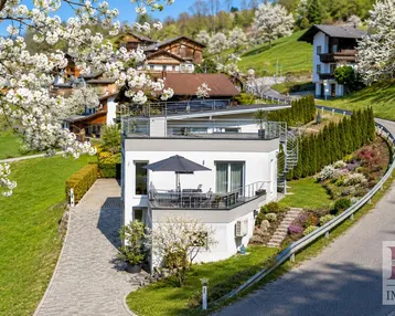 Modernes Haus mit mehreren Terrassen und Panoramablick in einer grünen Hügellandschaft mit blühenden Bäumen.