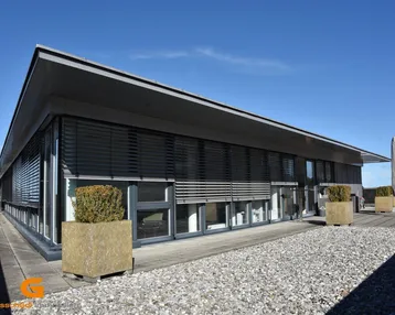 Moderne Bürogebäude-Fassade mit großen Fenstern, Jalousien und Kiesfläche im Außenbereich unter blauem Himmel.