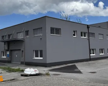 Modernes graues Gebäude mit vielen Fenstern und einem gepflasterten Vorplatz unter bewölktem Himmel.