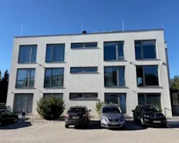 Modernes, weißes Gebäude mit großen Fenstern und Parkplätzen davor unter blauem Himmel.