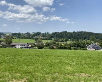 Weitläufige grüne Landschaft mit vereinzelten Häusern und bewaldeten Hügeln unter blauem Himmel.