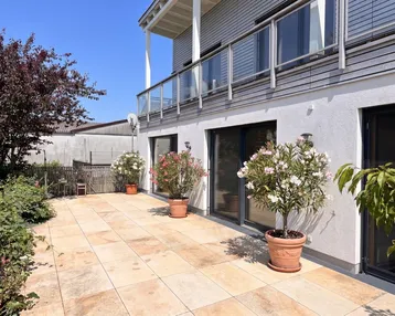 Große, sonnige Terrasse mit hellen Fliesen und blühenden Pflanzen vor der modernen Hausfassade.
