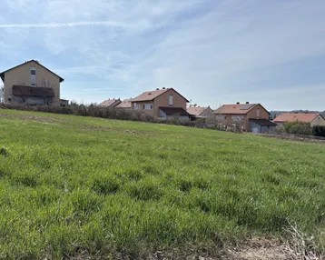 Weitläufiges grünes Feld mit Wohnhäusern im Hintergrund unter blauem Himmel.