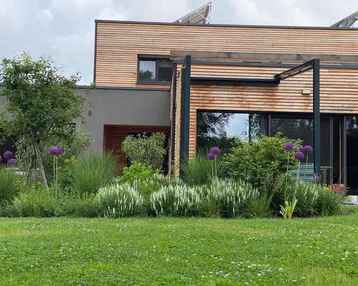Modernes Haus mit Holzfassade, großen Fenstern und einem gepflegten Garten mit vielfältiger Bepflanzung.