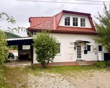 Charmantes Einfamilienhaus mit rotem Dach, weißer Fassade und überdachtem Außenbereich im Grünen.