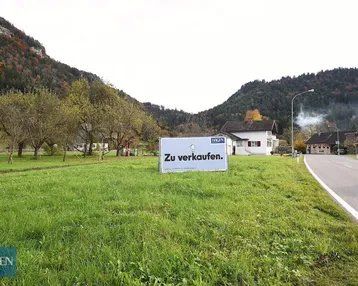 Grünes Grundstück mit 'Zu verkaufen'-Schild, umgeben von Bäumen und Bergen in ländlicher Umgebung.