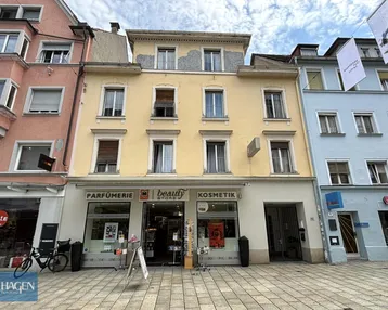 Gelbe Fassade eines Mehrfamilienhauses mit Geschäften im Erdgeschoss an einer belebten Straße.