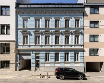 Historische Fassade eines Gebäudes in hellem Blau mit vielen Fenstern und einem geparkten Auto davor.