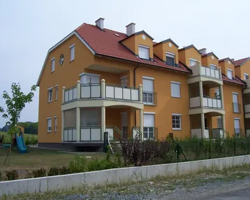 Mehrfamilienhaus mit gelber Fassade, rotem Ziegeldach und Balkonen, umgeben von Grünflächen und einem kleinen Spielplatz.