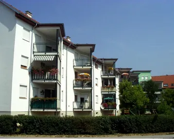 Modernes Mehrfamilienhaus mit weißen Fassaden, mehreren Balkonen, Markisen und grüner Umgebung.