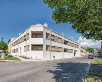Modernes weißes Mehrfamilienhaus an einer Straßenecke mit großen Fenstern, Außenjalousien und kleinen Grünflächen.