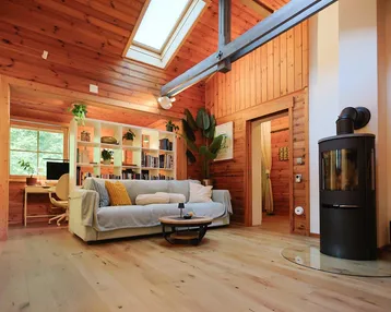 Gemütliches Wohnzimmer mit Holzwänden, Holzboden, Kaminofen und einem Arbeitsbereich unter dem Dachfenster.