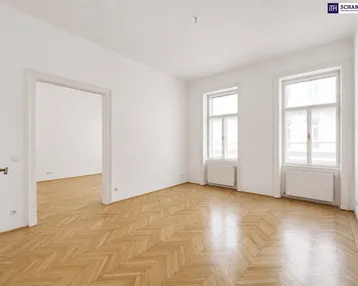Helles Zimmer mit Fischgrätparkett, hohen Decken und zwei großen Fenstern, offen zu einem weiteren Raum.