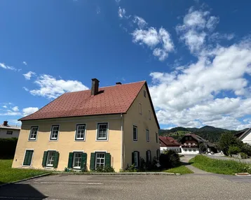 Traditionelles gelbes Haus mit rotem Ziegeldach und grünen Fensterläden unter blauem Himmel.