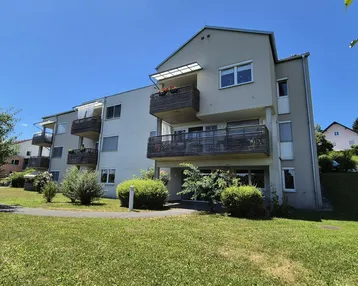 Modernes Mehrfamilienhaus mit mehreren Balkonen und gepflegter Grünfläche unter blauem Himmel.