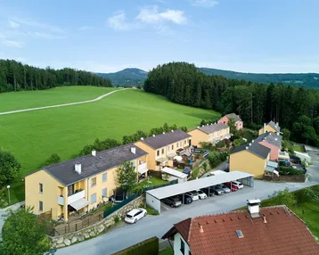 Luftaufnahme einer Wohnsiedlung mit Reihenhäusern, umgeben von grünen Feldern und Wald, inklusive Carport.
