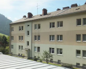Mehrfamilienhaus mit heller Fassade, Satteldach und Blick auf bewaldete Hügel.