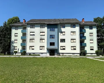 Mehrfamilienhaus mit hellgrüner Fassade und blauen Akzenten, umgeben von Grünflächen.