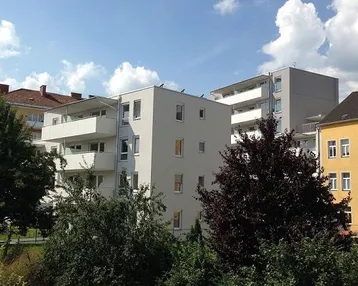 Modernes Mehrfamilienhaus mit hellgrauer Fassade, mehreren Balkonen und umgebender Grünfläche unter blauem Himmel.