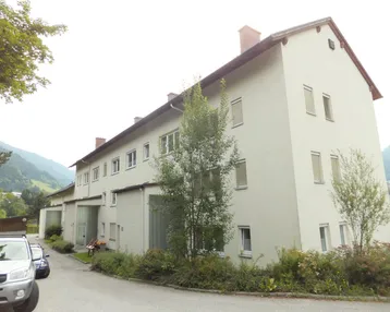 Gebäudeansicht mit heller Fassade, Satteldach und grüner Umgebung in einer ländlichen Gegend.