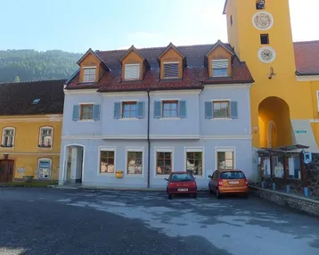 Gebäude mit hellblauer Fassade, Dachgauben und Blick auf die Berge, gelegen an einem Dorfplatz.