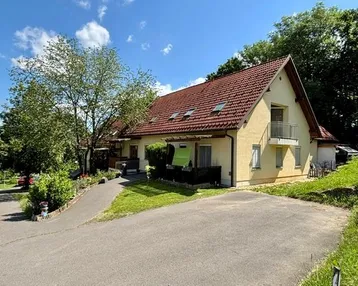 Gut gepflegtes Einfamilienhaus mit gelber Fassade, rotem Satteldach und kleinem Balkon, umgeben von viel Grün.