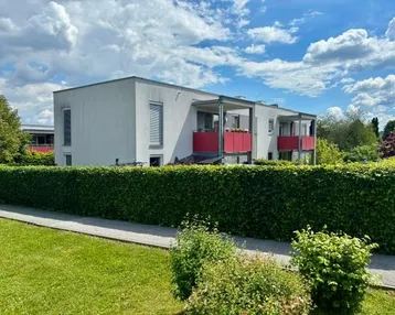 Modernes Gebäude mit weißer Fassade und roten Balkonen, umgeben von einer grünen Hecke unter blauem Himmel.
