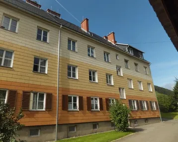 Mehrfamilienhaus mit gestreifter Fassade, traditionellen Fensterläden und grüner Umgebung unter blauem Himmel.