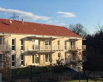 Mehrfamilienhaus mit mehreren Balkonen und angrenzender Grünfläche unter blauem Himmel.