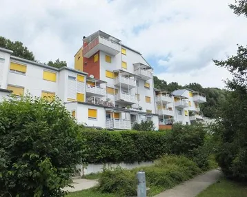 Mehrstöckiges Wohngebäude mit zahlreichen Balkonen und gelben Fensterläden, umgeben von viel Grün.