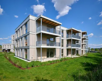 Modernes Mehrfamilienhaus mit hellen Fassaden und Balkonen, umgeben von grüner Landschaft unter blauem Himmel.