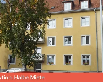 Gelbes Mehrfamilienhaus mit weißen Fenstern, einem kleinen Balkon und einem Baum im Vordergrund.