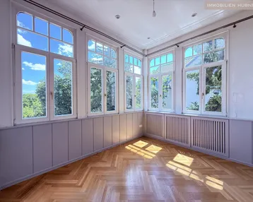 Helles Zimmer mit Fischgrätparkett und vielen Fenstern, die viel Tageslicht hereinlassen.