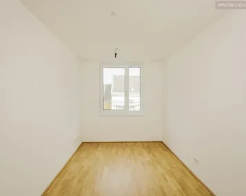 Leeres Zimmer mit hellem Parkettboden und einem Fenster, das viel Tageslicht hereinlässt.