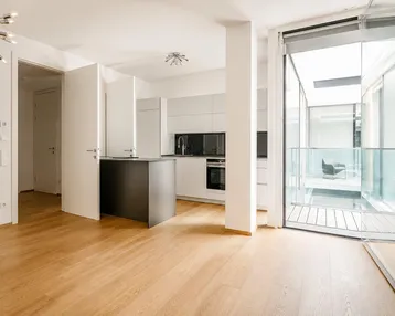 Heller offener Wohnbereich mit moderner Küche, Holzboden und Zugang zu einem verglasten Balkon.