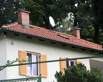 Dach des Hauses mit roten Ziegeln, Schornsteinen und Satellitenschüssel vor grünen Bäumen.