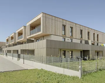 Moderne Wohnanlage mit Holzfassade und Balkonen, umgeben von Grünflächen unter blauem Himmel.