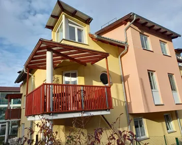 Fassade eines modernen Wohnhauses mit gelbem Putz, rotem Balkon und Dachgaube.