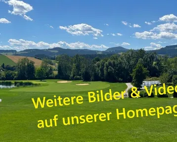Panoramablick auf einen Golfplatz mit Teich, umgeben von grünen Hügeln und Bergen unter blauem Himmel.