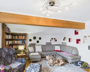 Gemütliches Wohnzimmer mit großem Sofa, Bücherregal, Holzbalken und moderner Deckenleuchte.
