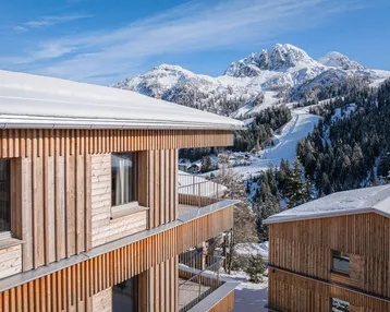Moderne Holzgebäude mit Balkonen vor einer verschneiten Berglandschaft und Skipisten.