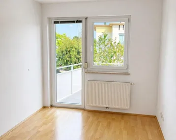 Helles Zimmer mit Parkettboden, großem Fenster und Zugang zu einem französischen Balkon.