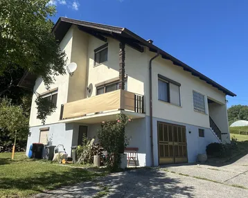 Einfamilienhaus mit Balkon und Gartenbereich in ländlicher Umgebung unter blauem Himmel.