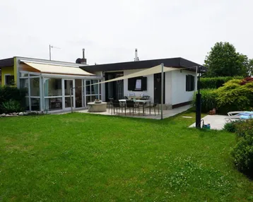 Haus mit großem Garten, überdachter Terrasse und Wintergarten, umgeben von viel Grün.