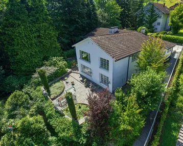 Luftaufnahme eines weißen Hauses mit Ziegeldach, umgeben von üppigem Grün und einem gepflasterten Gartenbereich.